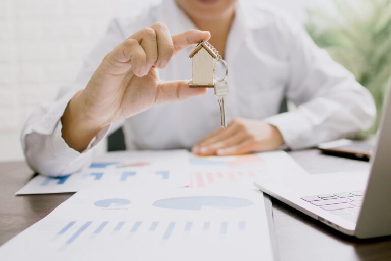 Hand holding a key on a small wooden house keyring above printed charts and a laptop